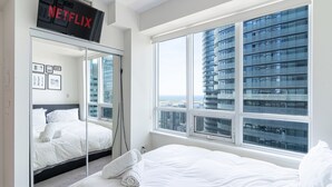 Ein modernes Hotelzimmer mit einem großen Bett, einem Kleiderschrank mit Spiegel, einem Fernseher, der 'NETFLIX' anzeigt, und einem großen Fenster mit Blick auf die Stadt.