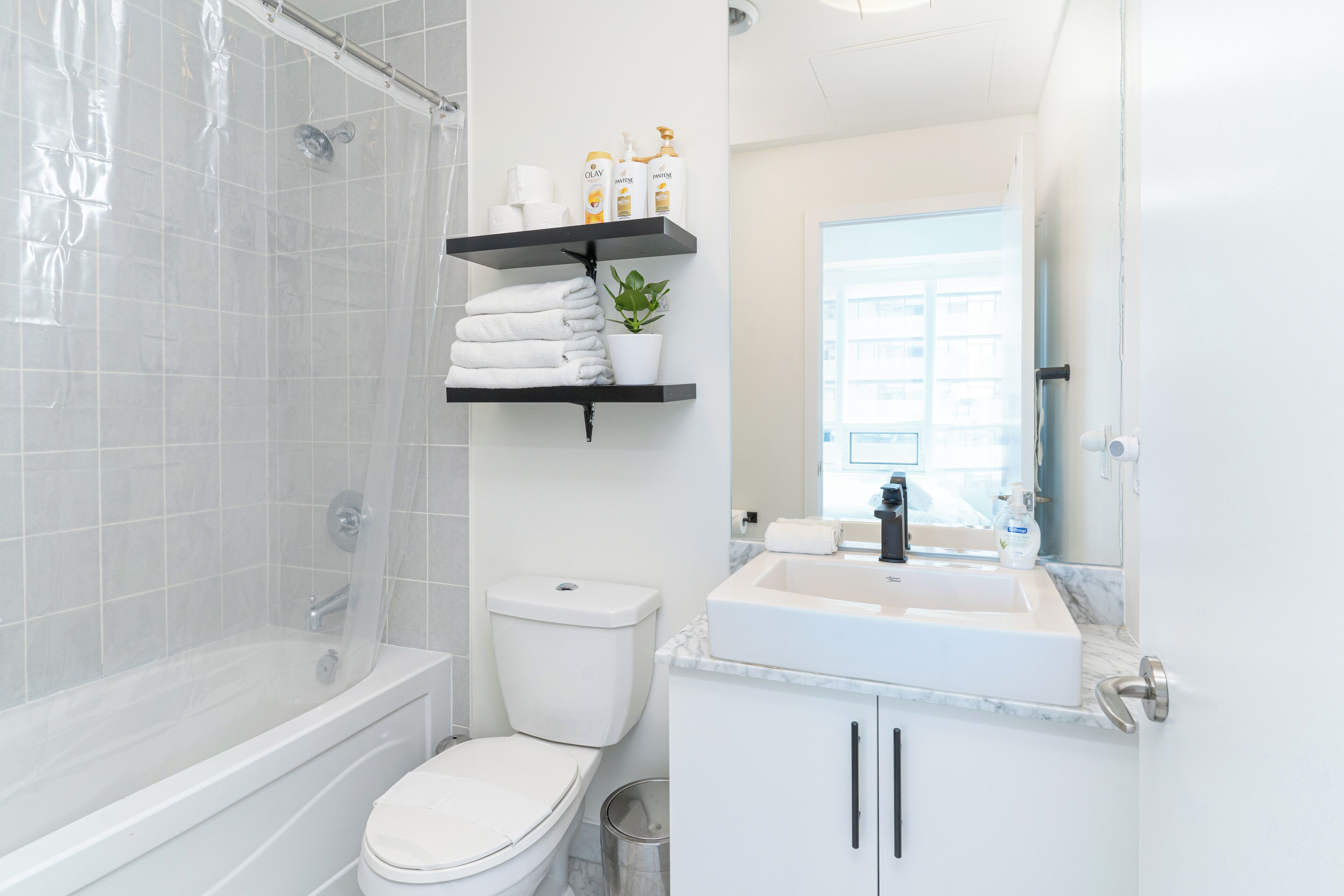 Et moderne badeværelse med badekar, toilet og håndvask med et spejl over. Der er hylder med toiletartikler og en potteplante.
