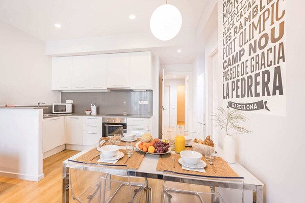 Una cocina moderna con armarios blancos, un microondas y un comedor con mesa de vidrio para dos.