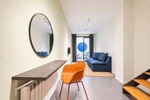 A modern living room with a sofa, a round mirror, and a dining area with a wooden table and orange chair.