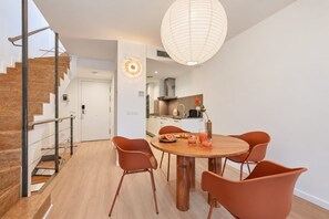 A modern interior with a dining area featuring a round wooden table and orange chairs, a kitchen with stainless steel appliances, and a staircase with wooden steps.