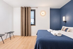 Une chambre à coucher avec un lit, une fenêtre avec des rideaux, une lampe fixée au mur et une petite table.