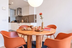 Un coin repas moderne avec une table ronde en bois, quatre chaises orange, un vase avec une fleur, un livre et une assiette de pain.