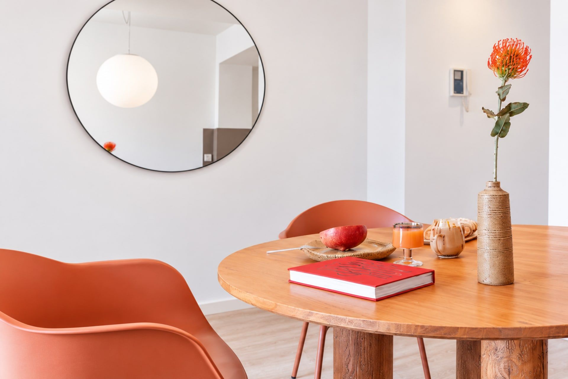Un comedor con una mesa redonda de madera, un libro rojo, un vaso de jugo de naranja y un florero con una flor.