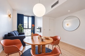 A modern living room with a round wooden dining table, orange chairs, a sofa, a large window with blue curtains, and a wall-mounted mirror. - Sweett | Girona 102 - One Bedroom Apartment, Sleeps 4 (Barcelona)