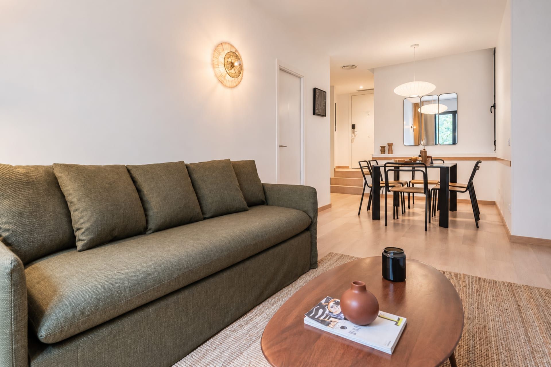 Una sala de estar con un sofá, una mesa de centro con una revista y un florero, un comedor con sillas y una mesa, y un pasillo con una puerta.