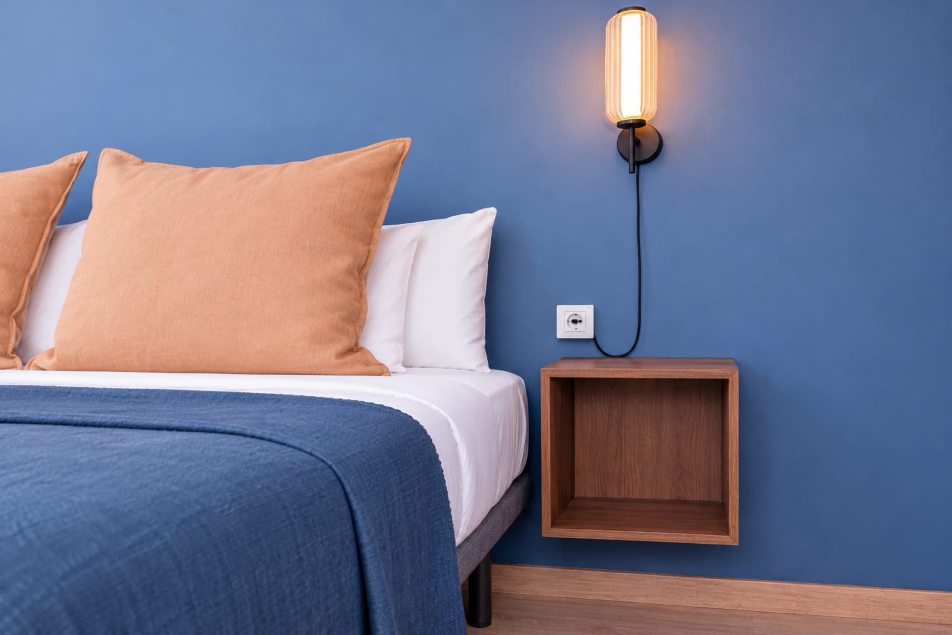 Una cama con colcha azul y dos almohadas, una lámpara de pared y una mesita de noche de madera con un estante.
