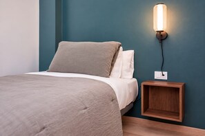 A neatly made bed with a grey duvet and pillows, a bedside table with a lamp, and a wall socket.
