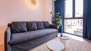 Una sala de estar moderna con un sofá gris, una mesita redonda, una lámpara de pie y un amplio ventanal con cortinas.