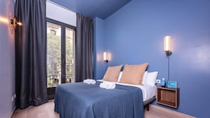 Ein Hotelzimmer mit einem großen Bett, zwei Nachttischlampen, einem Nachttisch mit einer Wasserflasche und Blick auf Gebäude durch das Fenster.