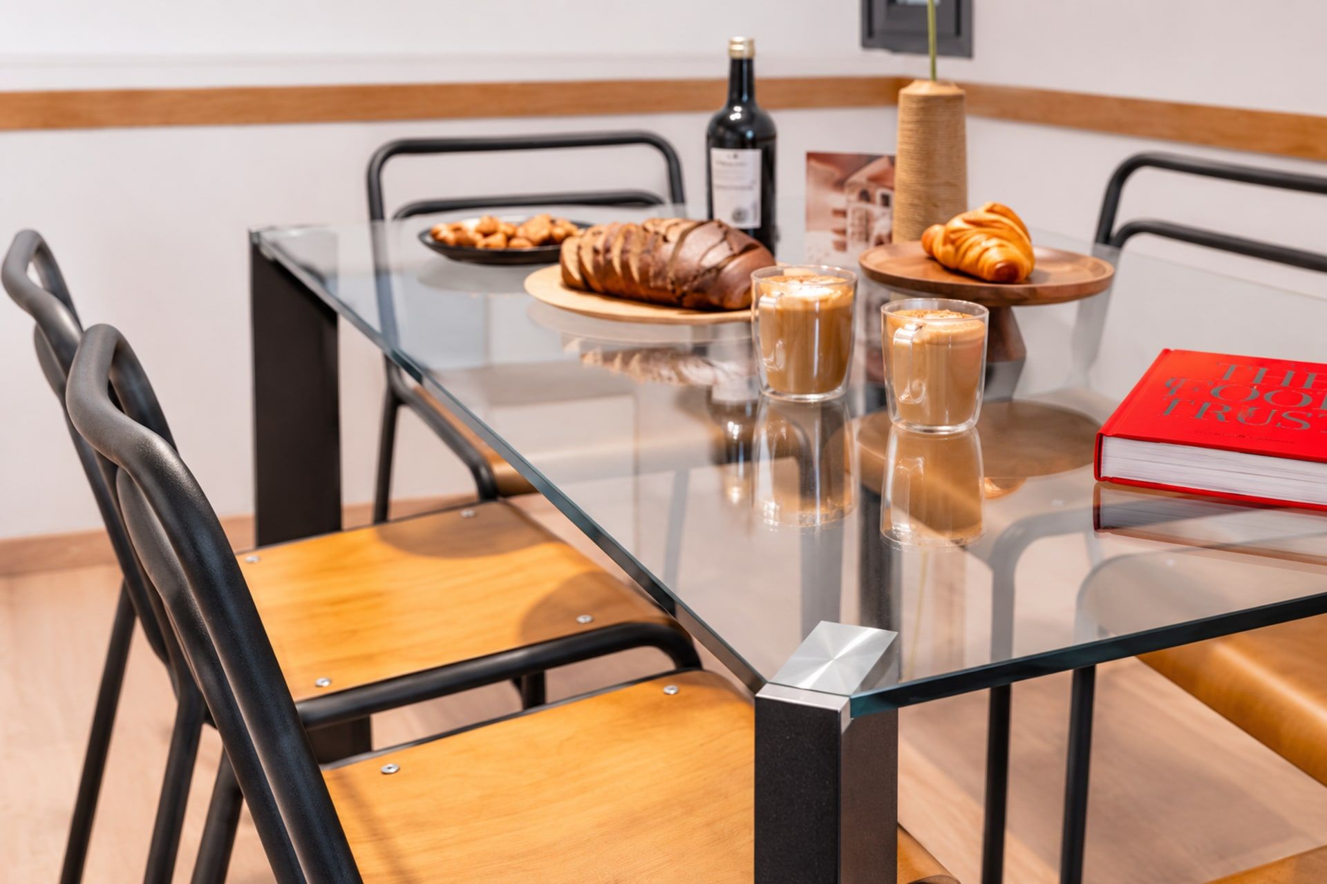 Una mesa con cubierta de vidrio, una botella de vino, croissants y tazas de café.