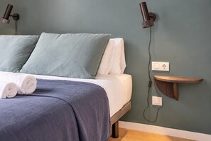 Ein ordentlich gemachtes Bett mit Kissen und Decke, ein Nachttisch mit Lampe und eine Steckdose an der Wand.