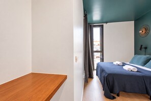 Une chambre à coucher avec un lit, un banc en bois, une fenêtre avec des rideaux et un ventilateur fixé au mur.