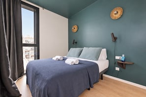A bedroom with a bed, a window with curtains, a wall-mounted lamp, and a small bedside table with a bottle.