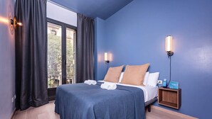 A hotel room with a large bed, two bedside lamps, a nightstand with a water bottle, and a view of buildings through the window. - Sweett | Girona 501 - Three Bedroom Apartment, Sleeps 7 (Barcelona)