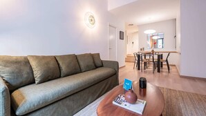 A living room with a sofa, a coffee table with a magazine, a vase, and a small plant. There is a dining area with a table and chairs in the background. - Sweett | Girona 501 - Three Bedroom Apartment, Sleeps 7 (Barcelona)