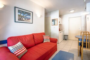 A red sofa with striped pillows, a framed picture on the wall, a dining table with a chair, and a kitchen area in the background.