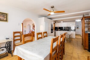 Een eetruimte met een tafel, stoelen, een plafondventilator en een keuken op de achtergrond.