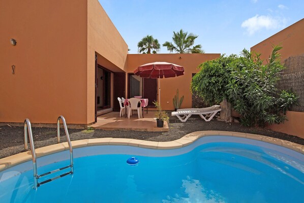 A swimming pool with a ladder, surrounded by a patio with chairs and an umbrella, and a cactus plant.