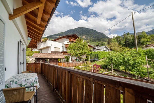 Un balcón de madera con una mesa y sillas, con vistas a un paisaje montañoso con casas y vegetación.