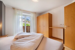 A bedroom with two beds, a wooden wardrobe, and a window with a view of a balcony and buildings.