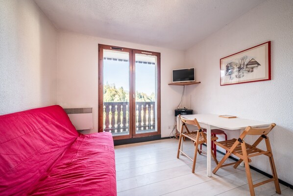 A room with a red couch, a white table with wooden chairs, a TV on a shelf, and a balcony with a view.