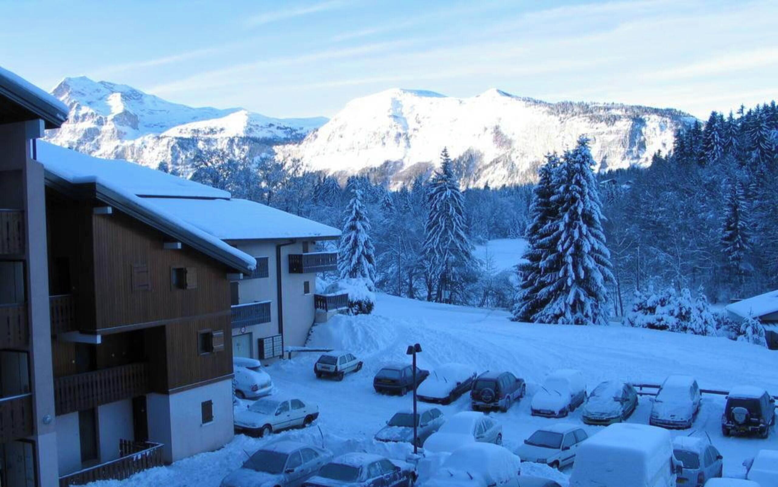 A building with a balcony, a parking lot with cars covered in snow, and snow-covered trees and mountains in the background.