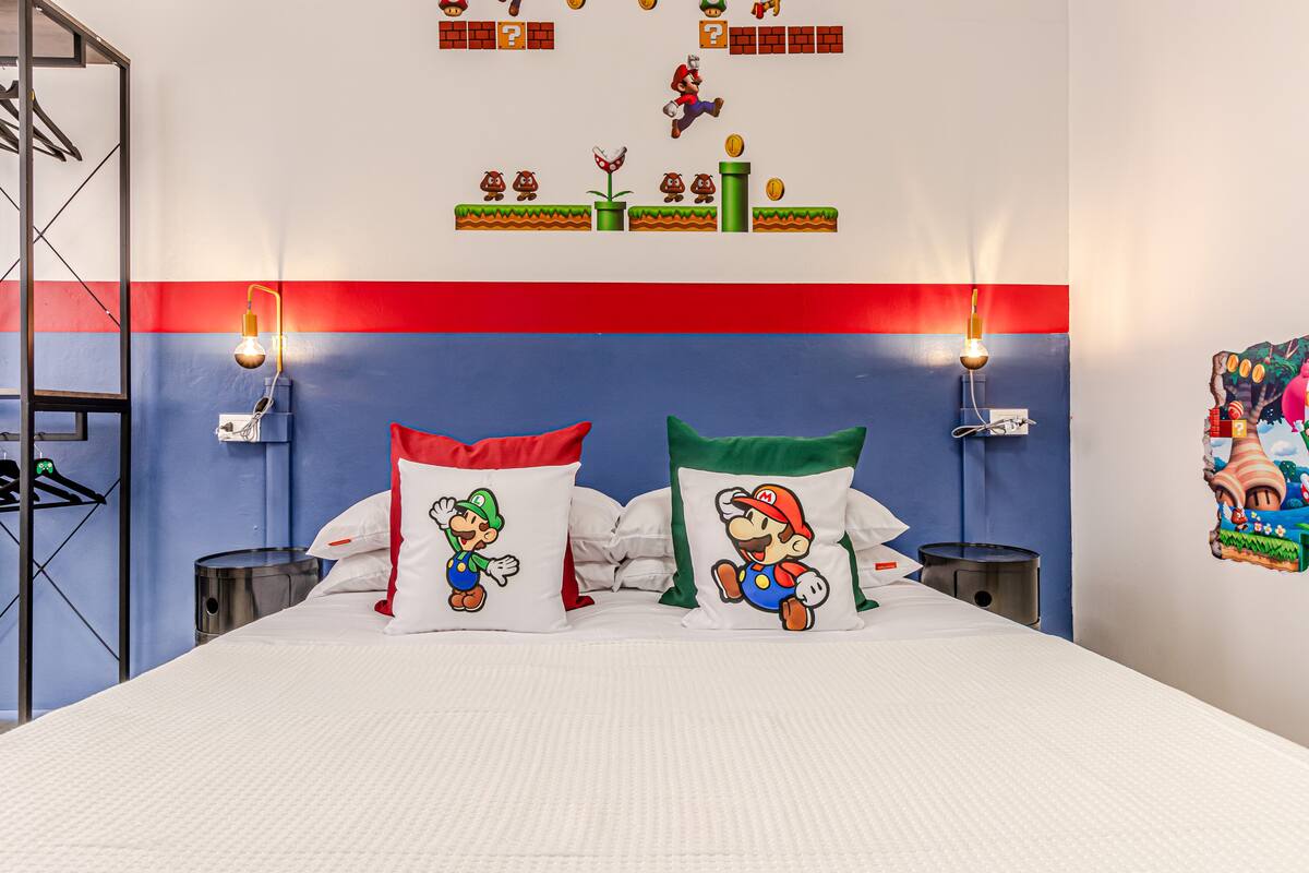 Habitación de hotel con una cama que presenta diseños de almohadas de Mario y Luigi, una pared a rayas rojas y azules, y una obra de arte con temática de Mario en la pared.