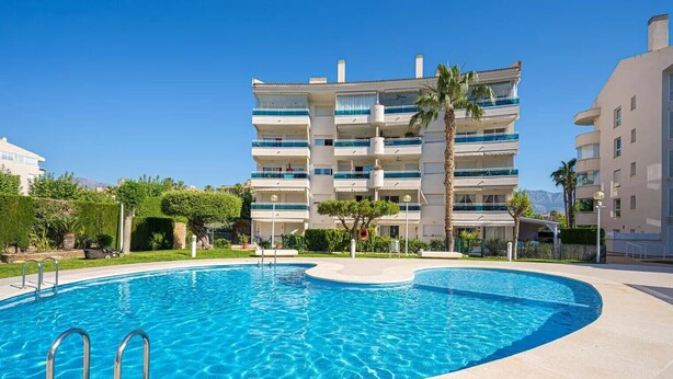 Una piscina de borde curvo, rodeada por un área ajardinada con palmeras y un edificio de varios pisos al fondo.