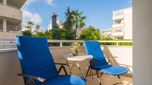 Dos sillones azules con una pequeña mesa y una planta en maceta entre ellos, ubicados en un balcón con vistas a palmeras y edificios.
