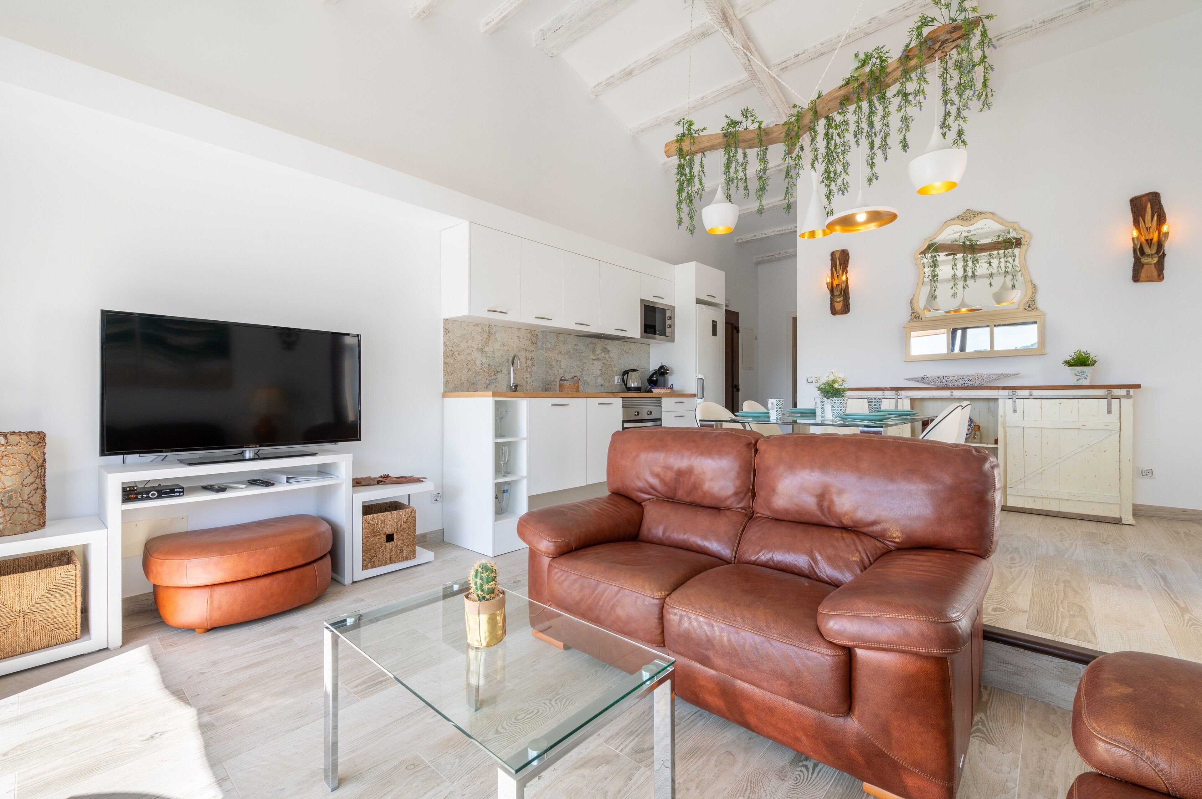 Una sala de estar moderna con un sofá de cuero, una mesa de centro de vidrio, un televisor de pantalla plana y una cocina al fondo.