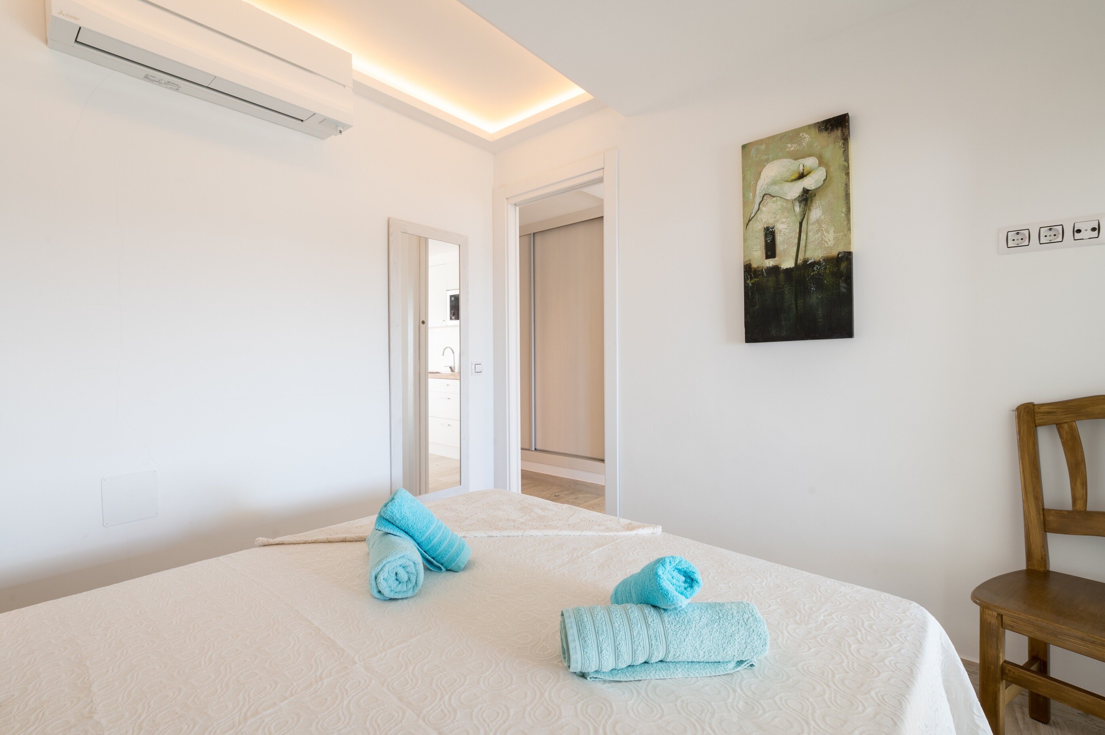 A hotel room with a bed, a chair, a painting, and an air conditioning unit.