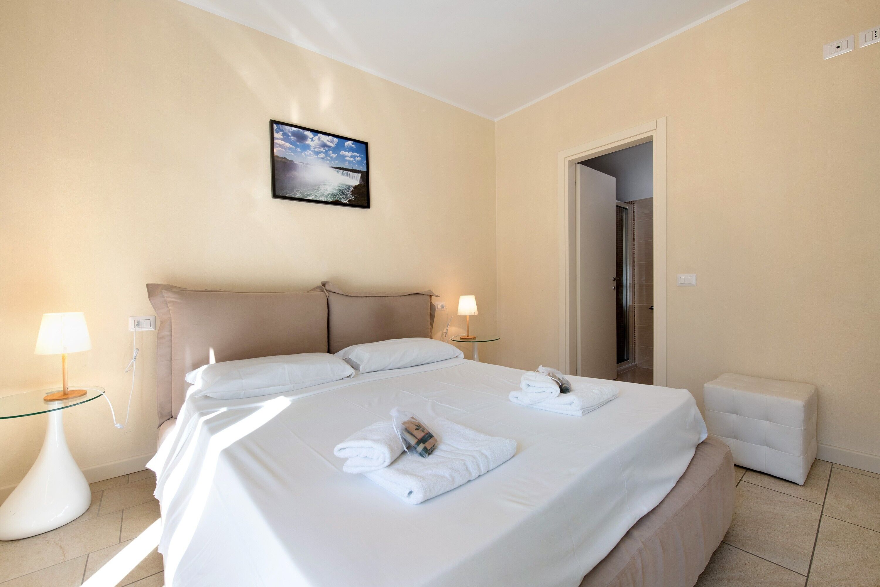 Une chambre d’hôtel avec un lit, deux lampes de chevet, une table de chevet, une petite table, un banc blanc et un tableau au mur.