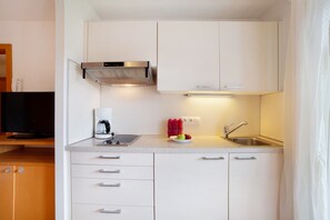 Une cuisine compacte avec des meubles blancs, une hotte de cuisinière en acier inoxydable, une machine à café, un évier et un petit panier à fruits.