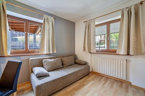 Una habitación con sofá, una silla, dos ventanas con cortinas, un radiador y techo de madera.