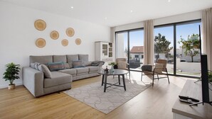 Uma sala de estar moderna com sofá, mesa de centro e poltronas, grandes janelas com cortinas e decoração na parede.
