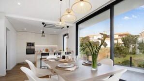 Cozinha moderna com armários brancos, área de refeições com mesa de madeira e uma janela grande com vista para edifícios e vegetação.