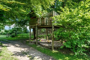 Une cabane en bois avec une échelle, entourée d’une végétation luxuriante et bordée d’un chemin de terre.