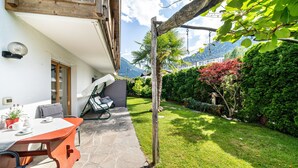 Un patio con una mesa y sillas, una pérgola de madera, un jardín cuidado con diversas plantas y vistas a montañas.