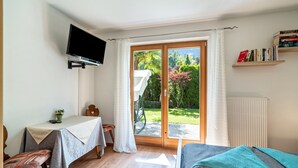 Un dormitorio con cama, una mesa con silla, televisor y vistas a un jardín a través de un ventanal corredera.