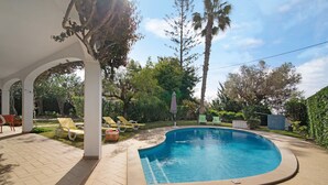 Ein Schwimmbecken mit gewölbter Kante, umgeben von einer überdachten Terrasse mit Stühlen und einem Tisch.