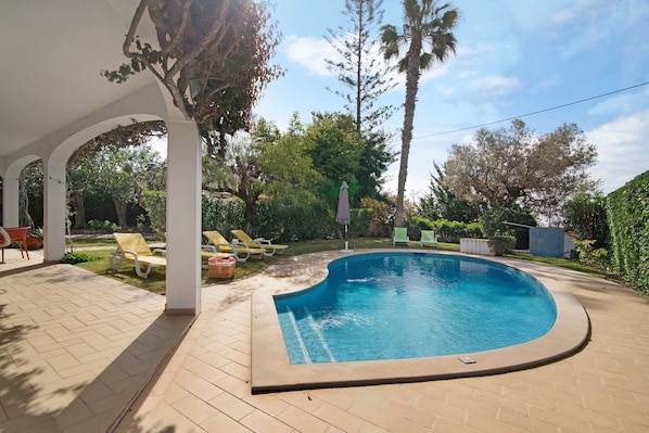 A swimming pool with a curved edge, surrounded by a covered patio area with chairs and a table.