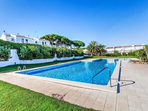 Une piscine rectangulaire, bordée d’une terrasse carrelée, entourée d’une pelouse et de haies bien entretenues, avec des bâtiments visibles au loin.