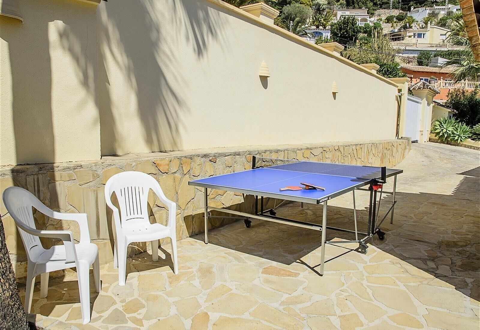 An outdoor table tennis setup with chairs, a stone patio, and a view of a hillside with buildings.