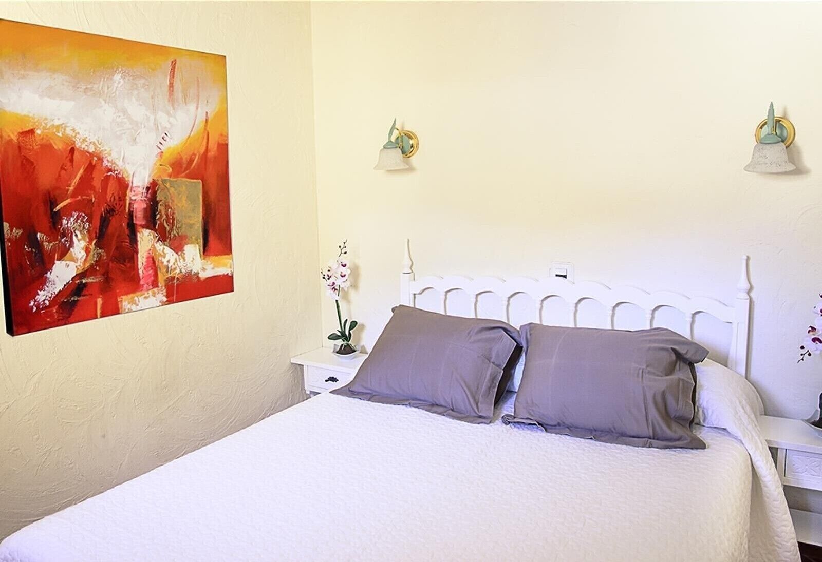 A bed with white headboard, two pillows, a nightstand with a lamp, and a painting on the wall.