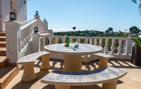 A round outdoor table with a tiled top, surrounded by four chairs, set on a balcony with a railing and a view of a residential area.