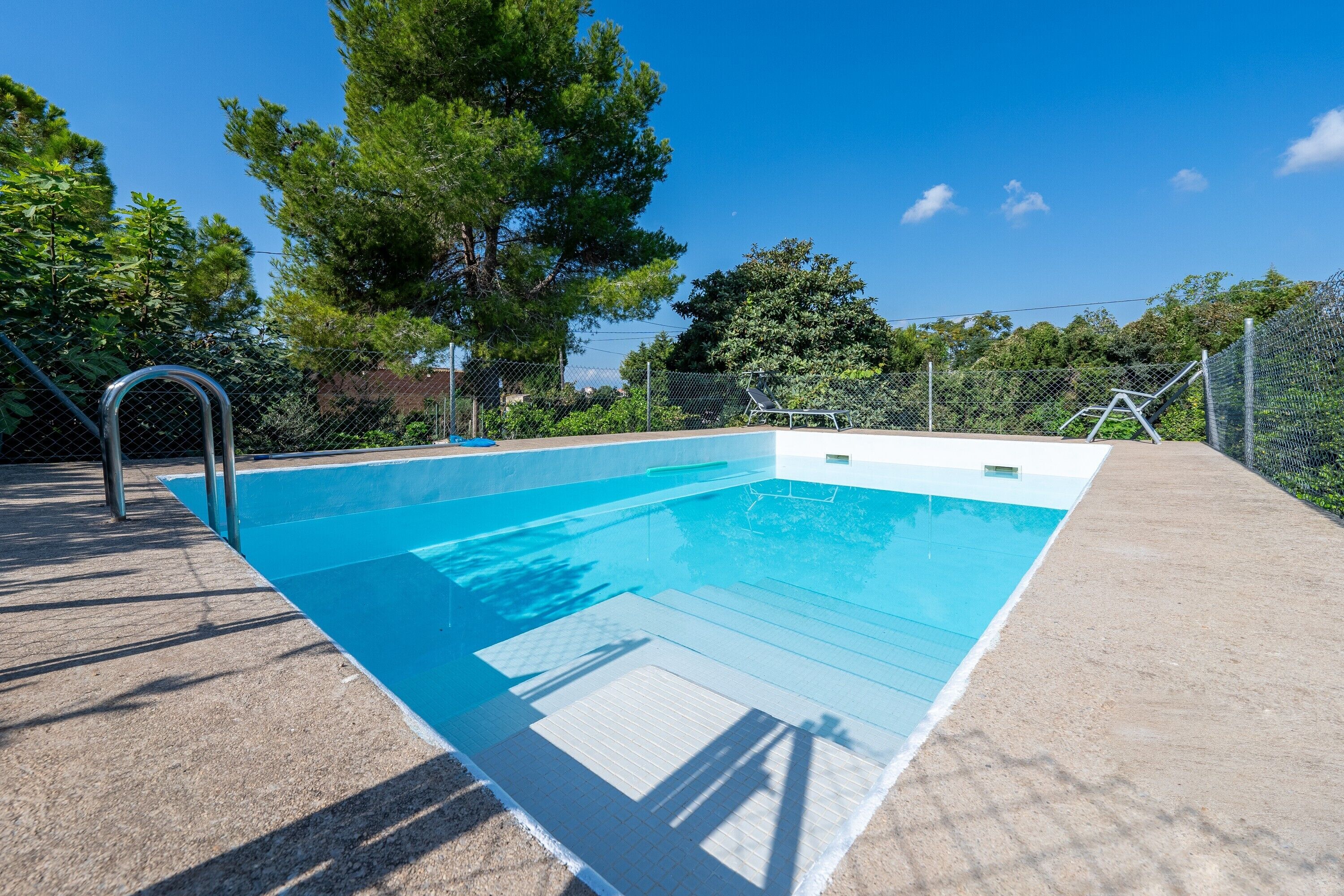 Une piscine rectangulaire avec un escalier en métal, entourée d’une terrasse en béton et d’une clôture en treillis métallique.