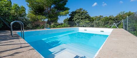 Une piscine rectangulaire avec un escalier en métal, entourée d’une terrasse en béton et d’une clôture en treillis métallique.