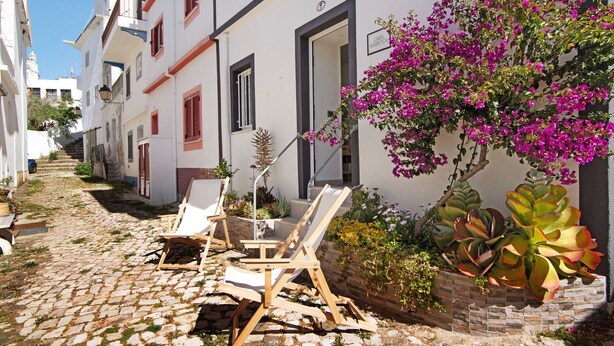 Una estrecha calle adoquinada con edificios blancos, un cartel que dice 'CASA BARROCO' y mesas y sillas para clientes al aire libre.
