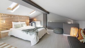 Un dormitorio moderno con techo inclinado, tragaluces, cama con cabecera de madera, mesita de noche y sofá gris con cojines estampados.